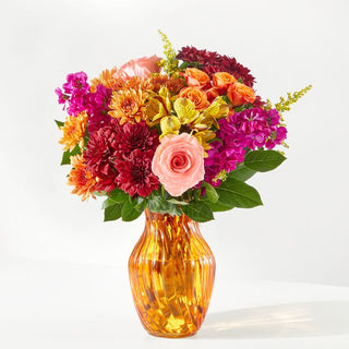 Vibrant fall flower arrangement with orange roses, coral pink roses, burgundy chrysanthemums, and fuchsia stock in an amber glass vase — autumn bouquet Toronto florist.