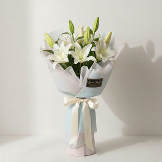 A hand-wrapped floral bouquet from Yunza Florist featuring three stems of pristine white lilies with several closed green buds. The arrangement is styled in a modern Korean-style wrap using layered, pale blue and translucent white matte paper, finished with a cream satin ribbon.