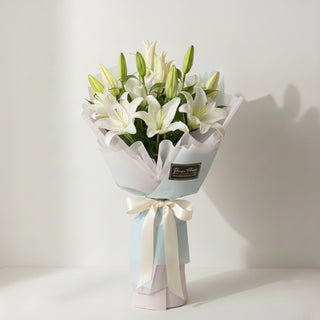 A large, premium bouquet of white lilies by Yunza Florist, showing a lush arrangement of six or more stems with multiple open blooms and fresh green buds. The bouquet is professionally wrapped in voluminous, layered Korean-style decorative paper in soft pastel tones and tied with a wide cream ribbon, showcasing a full and luxurious aesthetic.